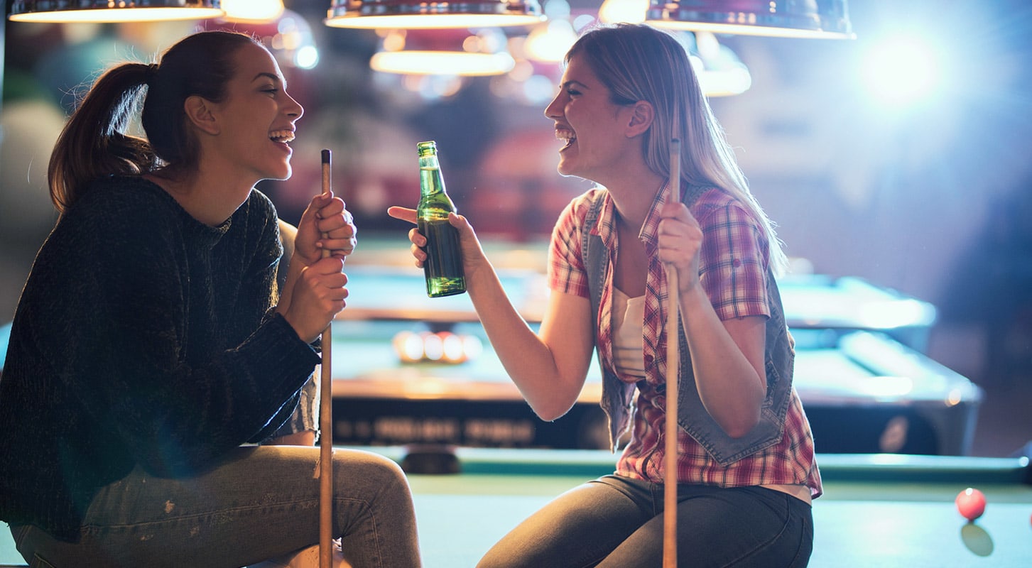 Two women sitting on a pool table, enjoying beers and laughing together in a casual setting.